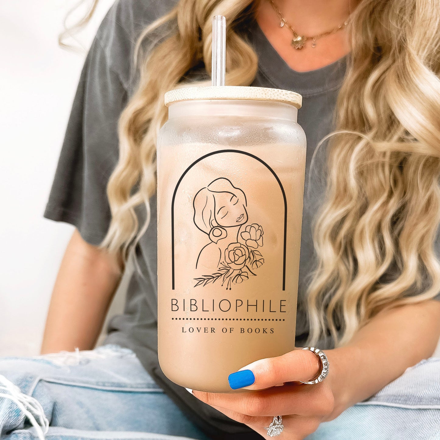 A frosted glass cup with a bamboo lid and glass straw featuring a minimalist line art illustration of a woman holding flowers, accompanied by the words "Bibliophile - Lover of Books." The cup is held by a woman with long, wavy blonde hair, wearing a casual grey t-shirt and blue nail polish. Perfect for book lovers, this aesthetic bookish glass cup is ideal for iced coffee, tea, or any favorite beverage. A must-have for readers who adore stylish and literary-inspired drinkware.