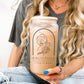 A frosted glass cup with a bamboo lid and glass straw featuring a minimalist line art illustration of a woman holding flowers, accompanied by the words "Bibliophile - Lover of Books." The cup is held by a woman with long, wavy blonde hair, wearing a casual grey t-shirt and blue nail polish. Perfect for book lovers, this aesthetic bookish glass cup is ideal for iced coffee, tea, or any favorite beverage. A must-have for readers who adore stylish and literary-inspired drinkware.